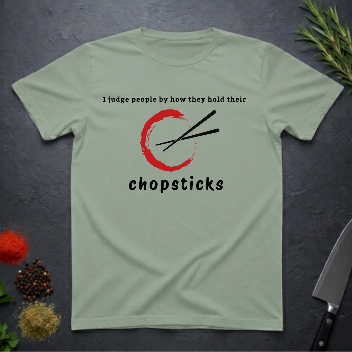 Sage green t-shirt with Chopsticks graphic and text "I judge people by how they hold their chopsticks" on a dark surface with kitchen items.