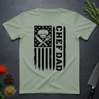 Sage green t-shirt with American flag and text 'CHEF DAD' design on a dark surface with kitchen items.
