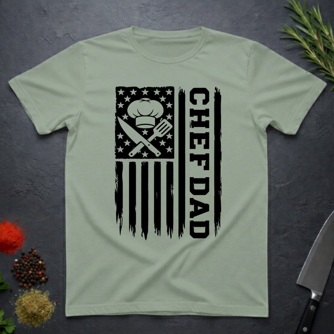 Sage green t-shirt with American flag and text 'CHEF DAD' design on a dark surface with kitchen items.
