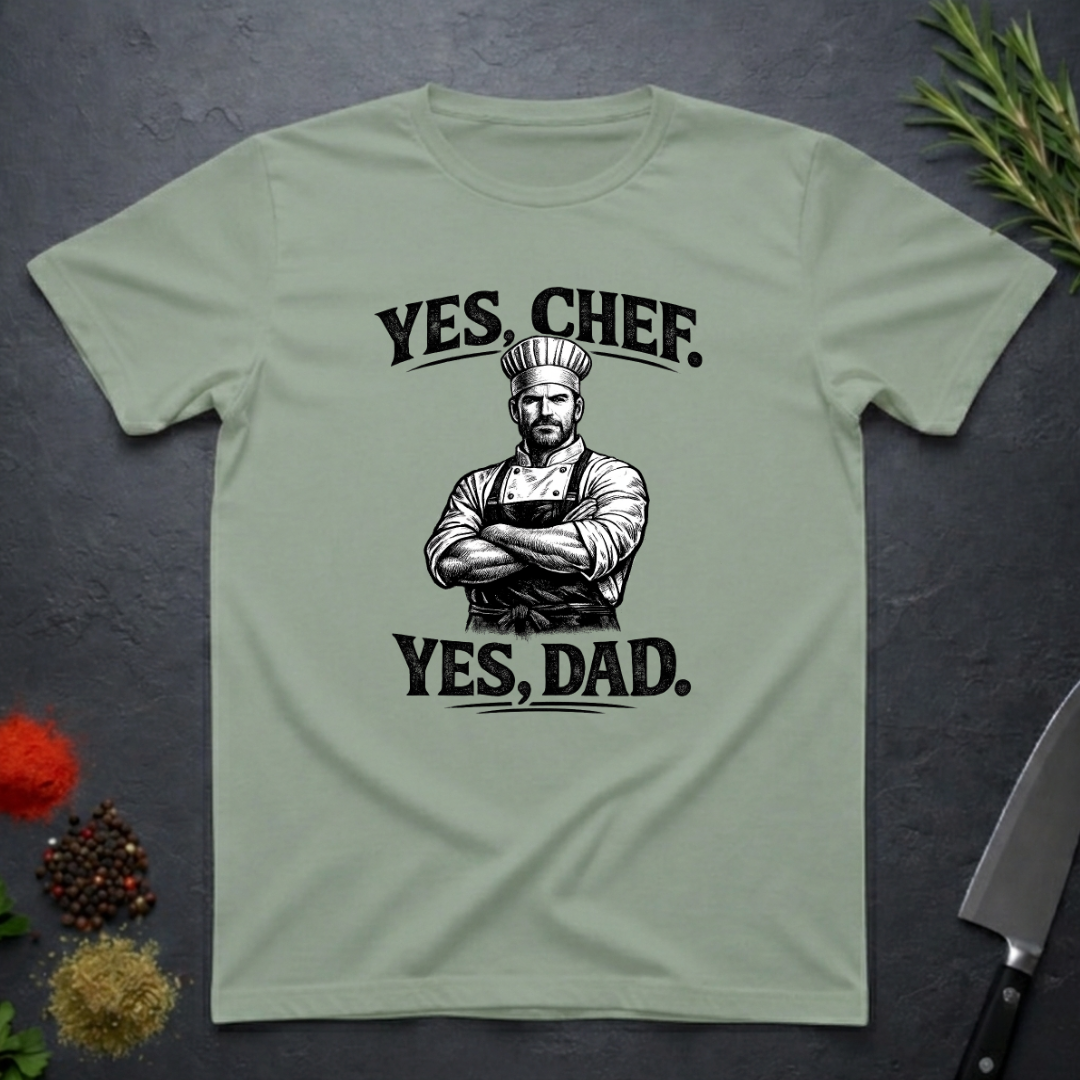 Sage green t-shirt with a graphic of a chef and text 'YES, CHEF. YES, DAD.' on a dark surface.