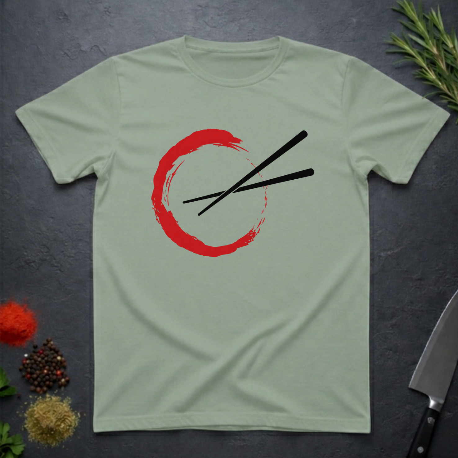 Sage green t-shirt with a red circle and black chopsticks design on a dark surface.