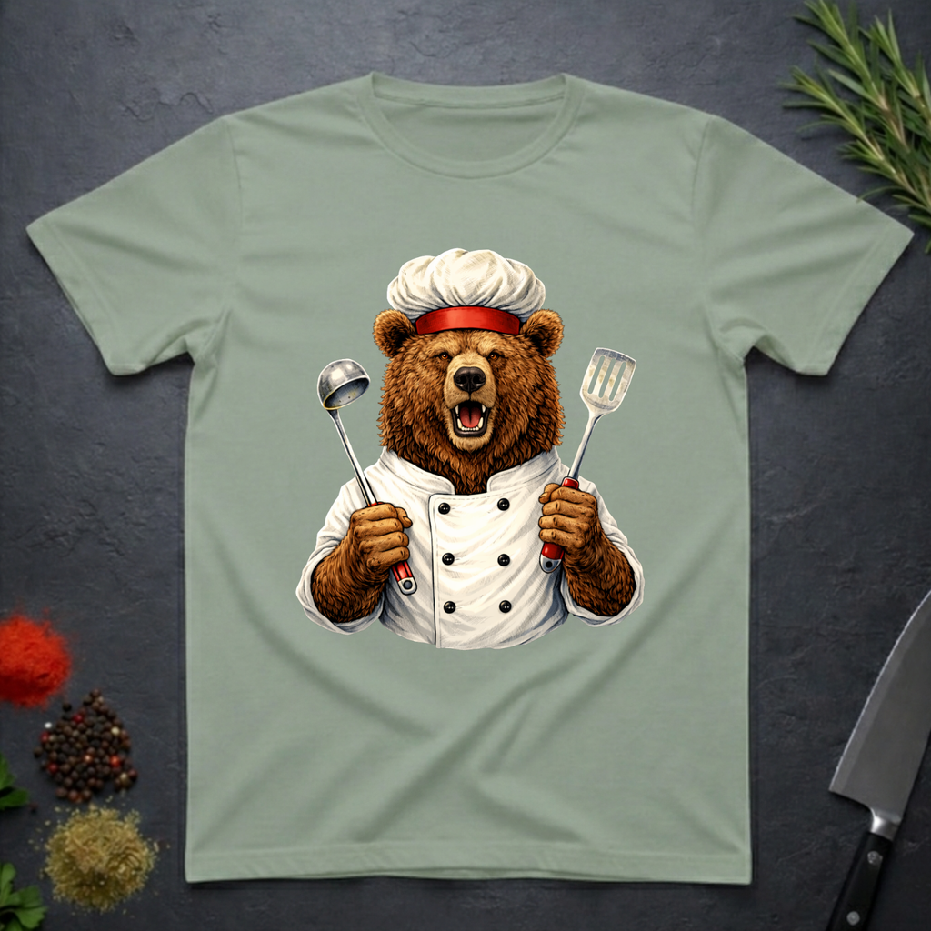 Sage green t-shirt with a grizzly bear chef holding utensils design on a dark surface.