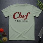 Sage green t-shirt with 'Chef A Title Earned' text on a dark surface with kitchen items.