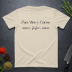 Sand colored t-shirt with Spanish text "Pan, Vino y Carne" on a dark surface with decorative elements. 