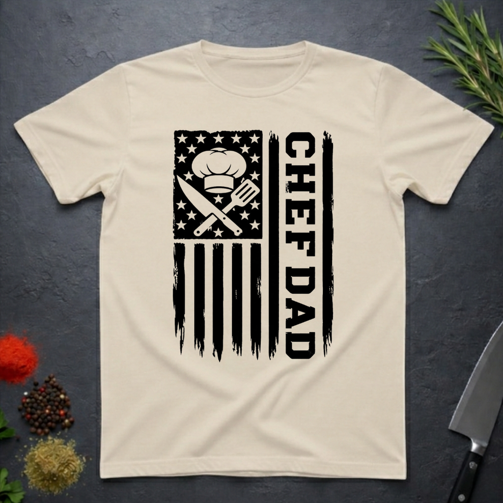 Sand colored t-shirt with American flag and text 'CHEF DAD' design on a dark surface with kitchen items.