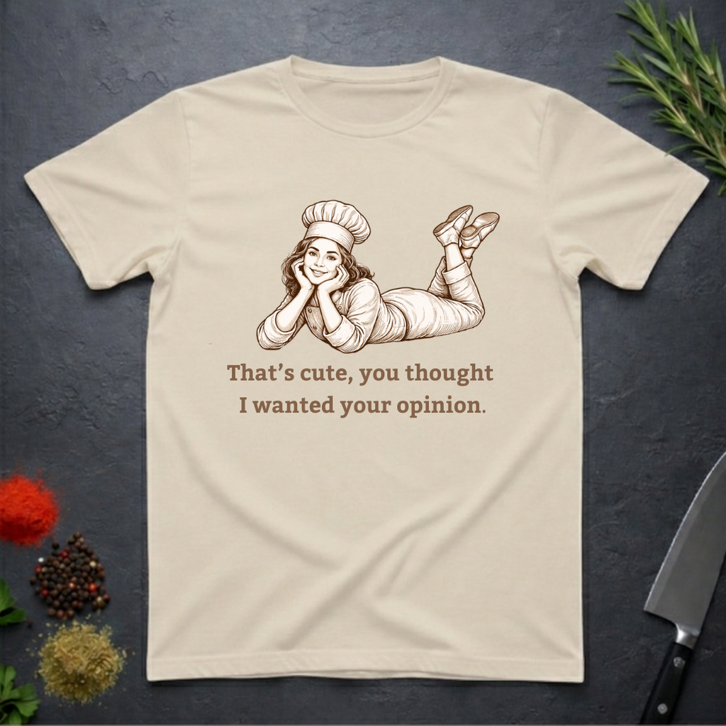 Sand colored t-shirt with a graphic of a woman in a chef's outfit and text "That's cute, you thought I wanted your opinion." On a dark surface.