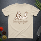 Sand colored t-shirt with a graphic of a woman in a chef's outfit and text "That's cute, you thought I wanted your opinion." On a dark surface.