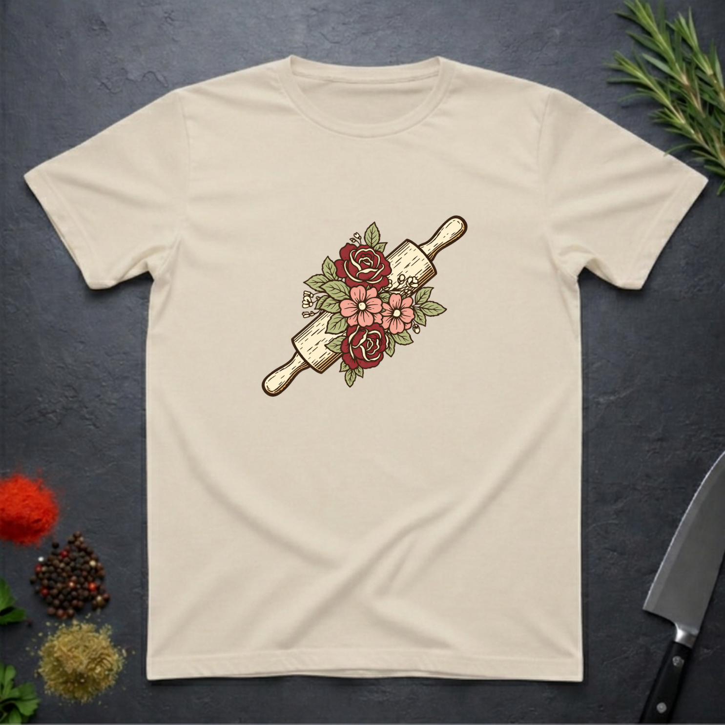 Sand colored t-shirt with a rolling pin and floral design on a dark surface.