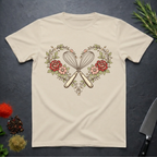 Sand colored t-shirt with a design of crossed kitchen utensils and flowers on a dark surface.