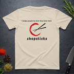 Sand colored t-shirt with Chopsticks graphic and text "I judge people by how they hold their chopsticks" on a dark surface with kitchen items.