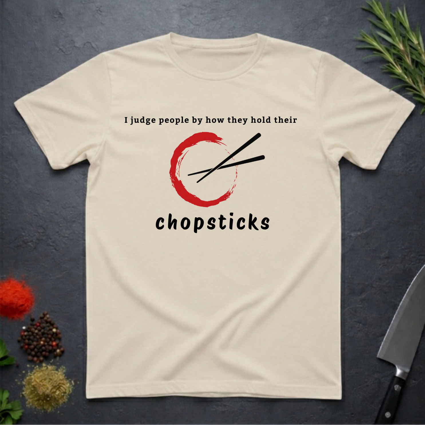 Sand colored t-shirt with Chopsticks graphic and text "I judge people by how they hold their chopsticks" on a dark surface with kitchen items.