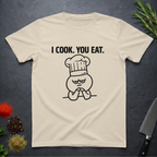 Sand colored t-shirt with a graphic design and text 'I COOK. YOU EAT.' on a dark surface with kitchen items.