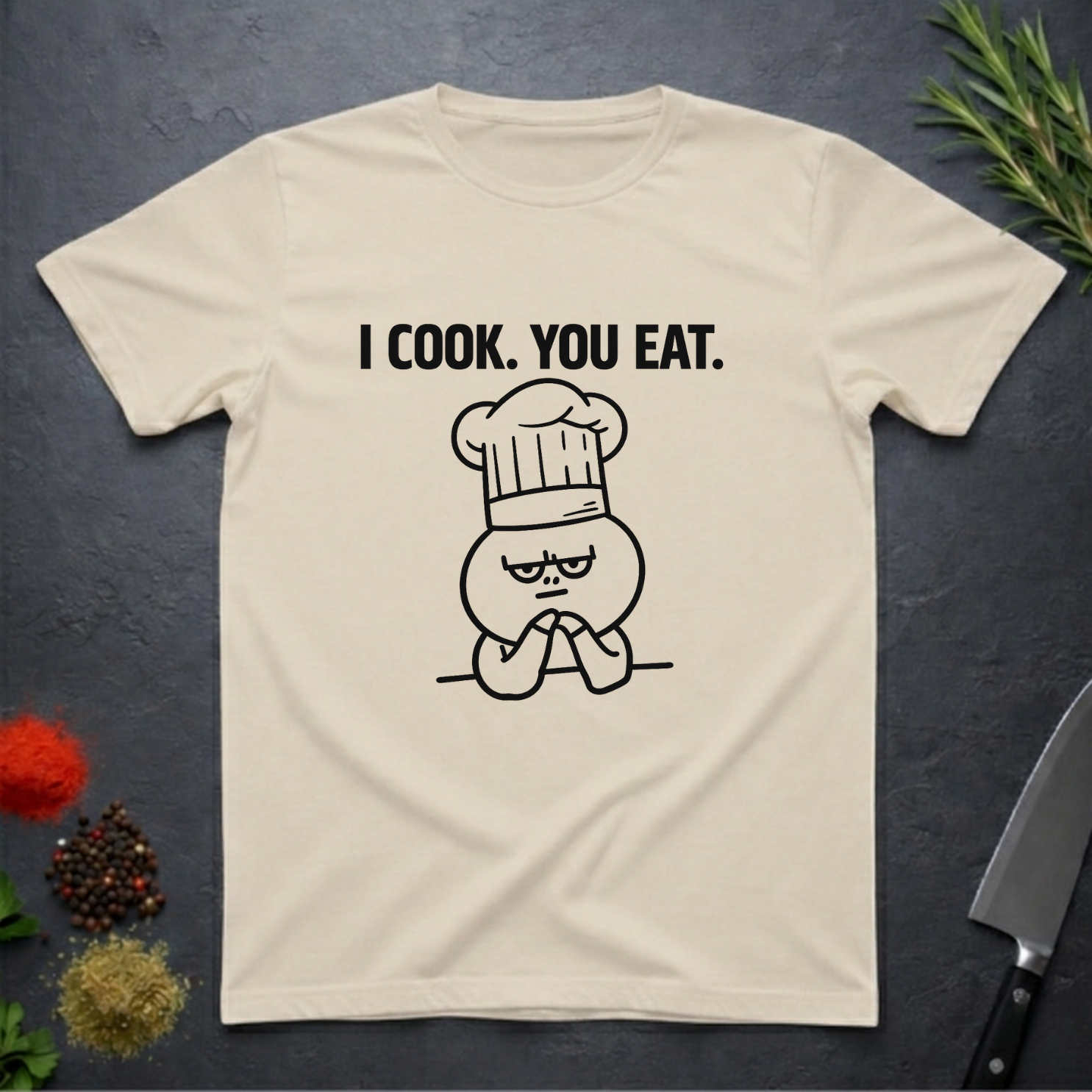 Sand colored t-shirt with a graphic design and text 'I COOK. YOU EAT.' on a dark surface with kitchen items.