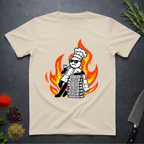 Sand colored t-shirt with a cartoon dead-pan chef leaning on text books with a background of flames on a dark surface.