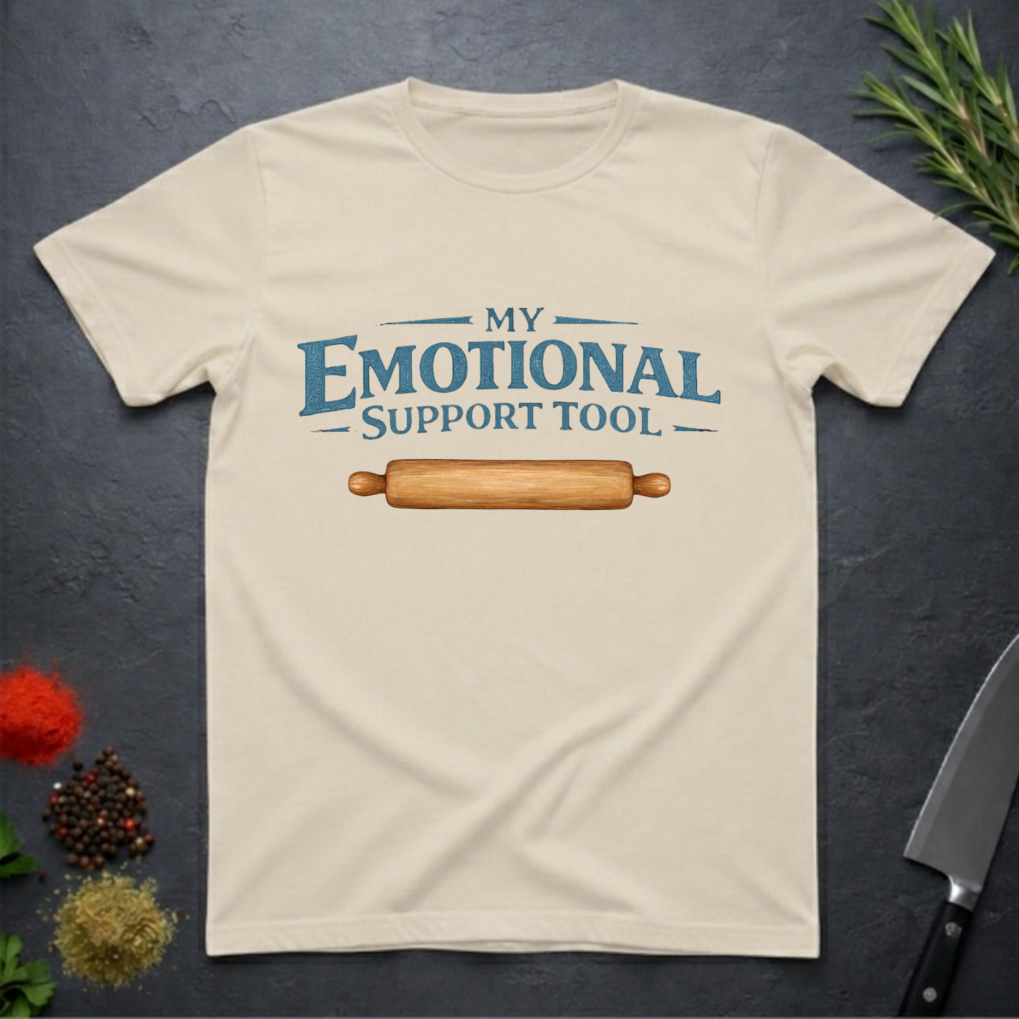 Sand colored t-shirt with a graphic of a rolling pin and text 'My Emotional Support Tool' on a dark surface.