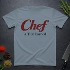 Stone blue t-shirt with 'Chef A Title Earned' text on a dark surface with kitchen items.