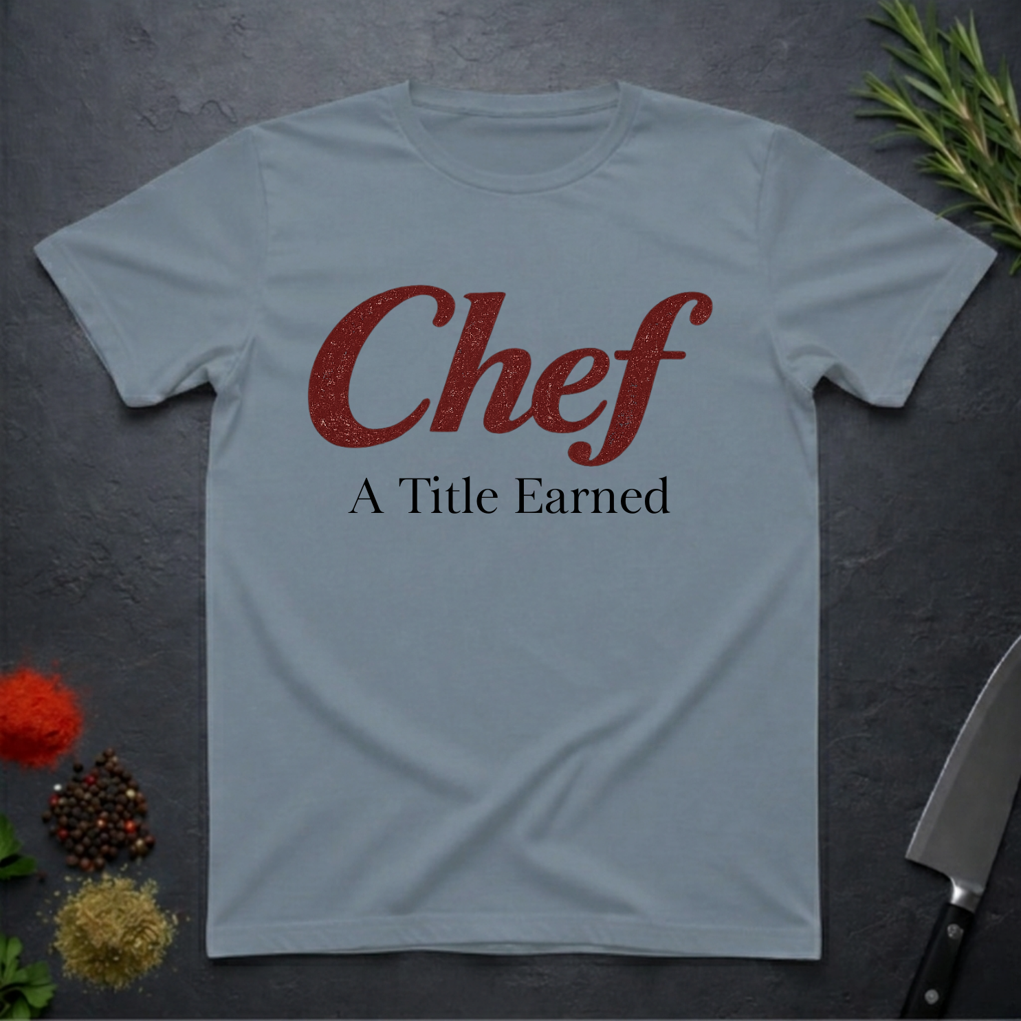 Stone blue t-shirt with 'Chef A Title Earned' text on a dark surface with kitchen items.