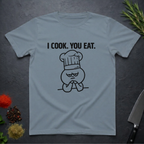 Stone blue t-shirt with a graphic and text 'I COOK. YOU EAT.' on a dark surface with kitchen items.