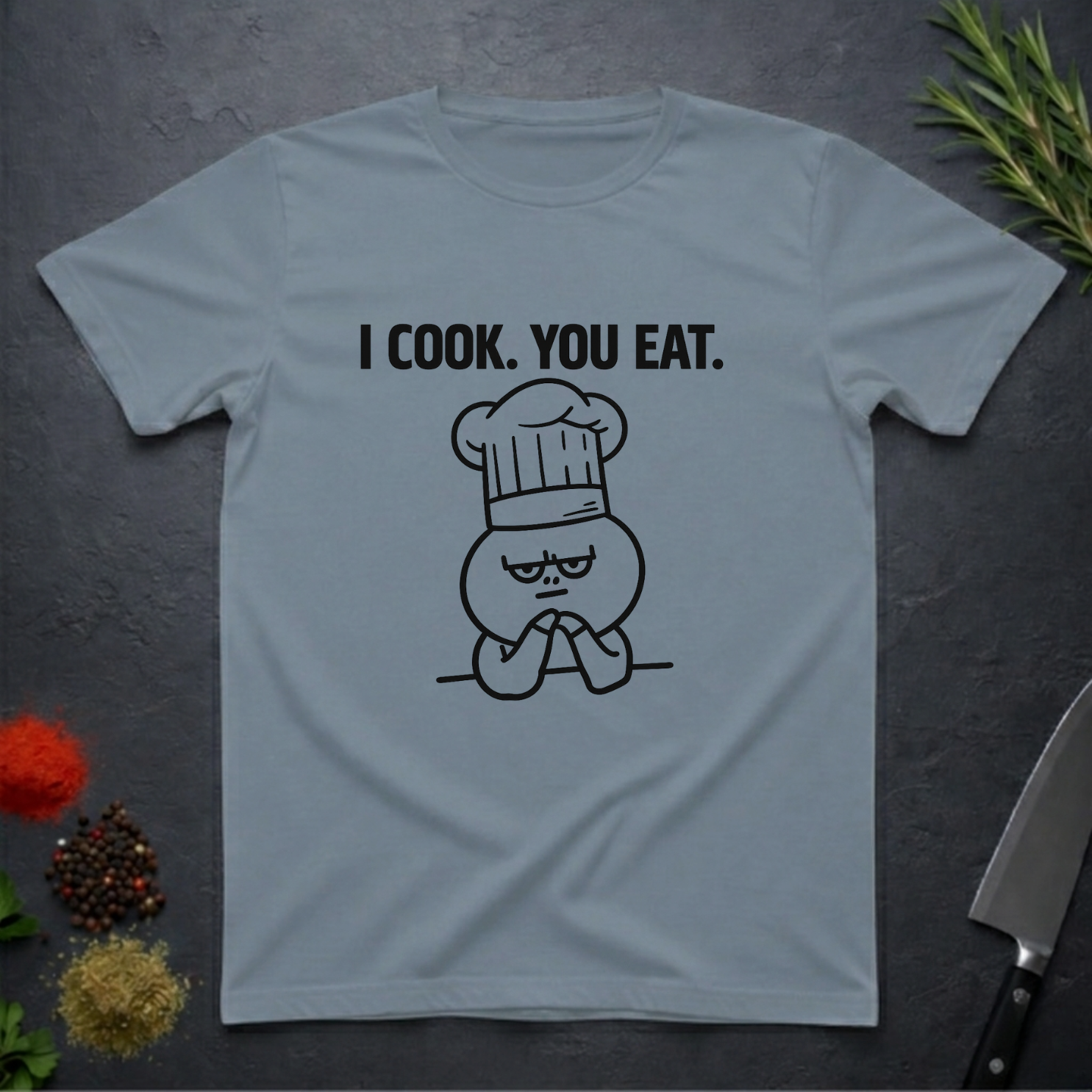 Stone blue t-shirt with a graphic and text 'I COOK. YOU EAT.' on a dark surface with kitchen items.