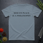 Stone blue t-shirt with 'MISE EN PLACE IS A PHILOSOPHY' text on a dark surface with kitchen items.