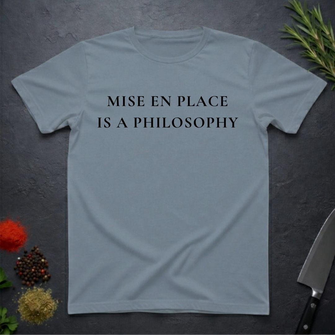 Stone blue t-shirt with 'MISE EN PLACE IS A PHILOSOPHY' text on a dark surface with kitchen items.