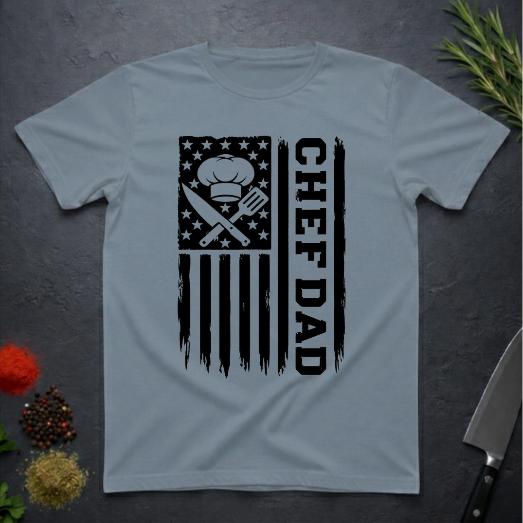 Stone blue t-shirt with American flag and text 'CHEF DAD' design on a dark surface with kitchen items.