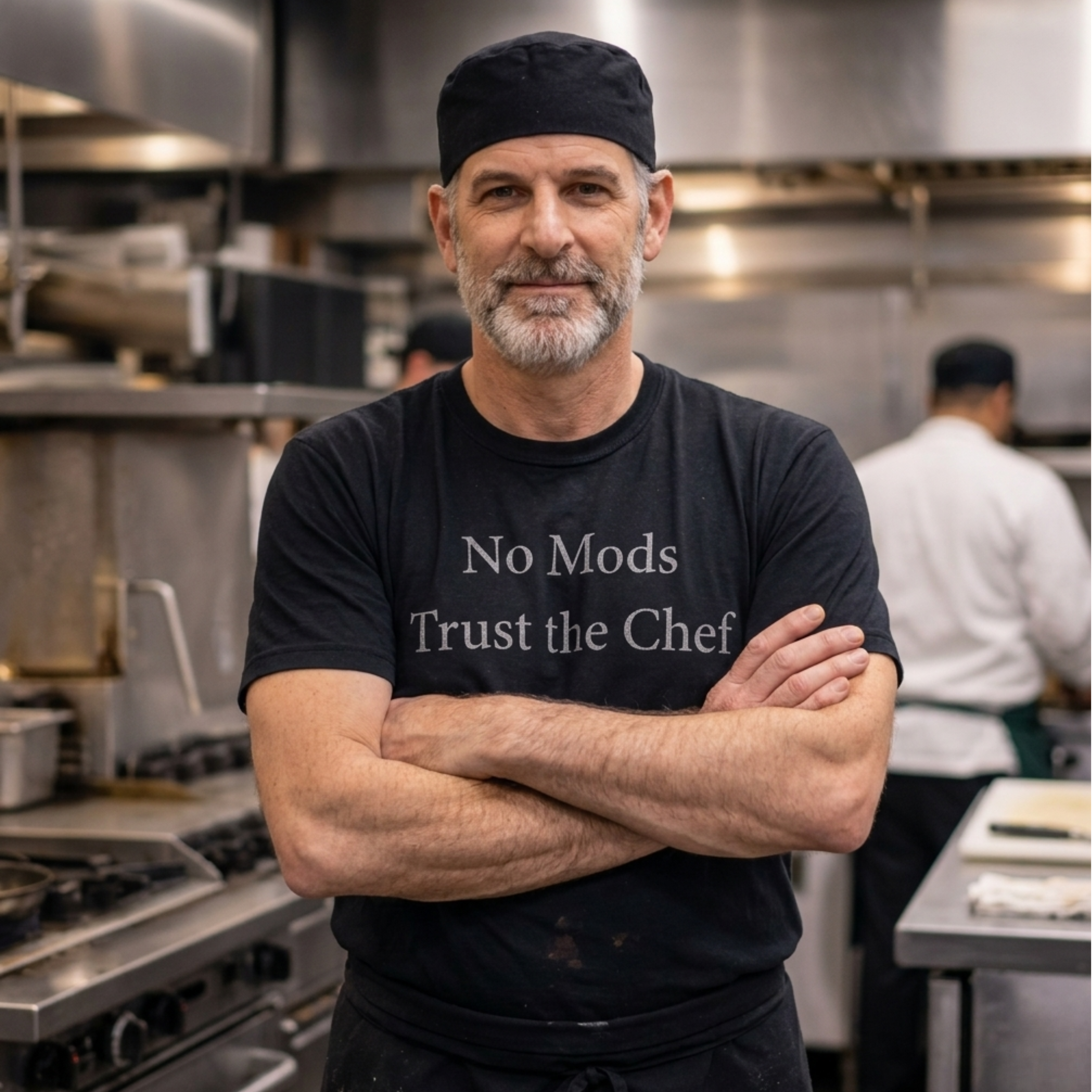 Older Male Chef in a kitchen wearing a black t-shirt with text "No Mons Trust the Chef", standing with arms crossed.
