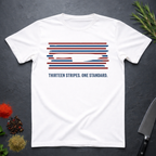 White t-shirt with 13 red, white, and blue stripes with a cut out (negative space) image of a chef's knife and text "THIRTEEN STRIPES. ONE STANDARD." on a dark surface.