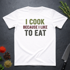 White t-shirt with 'I cook because I like to eat' text on a dark surface with kitchen items.