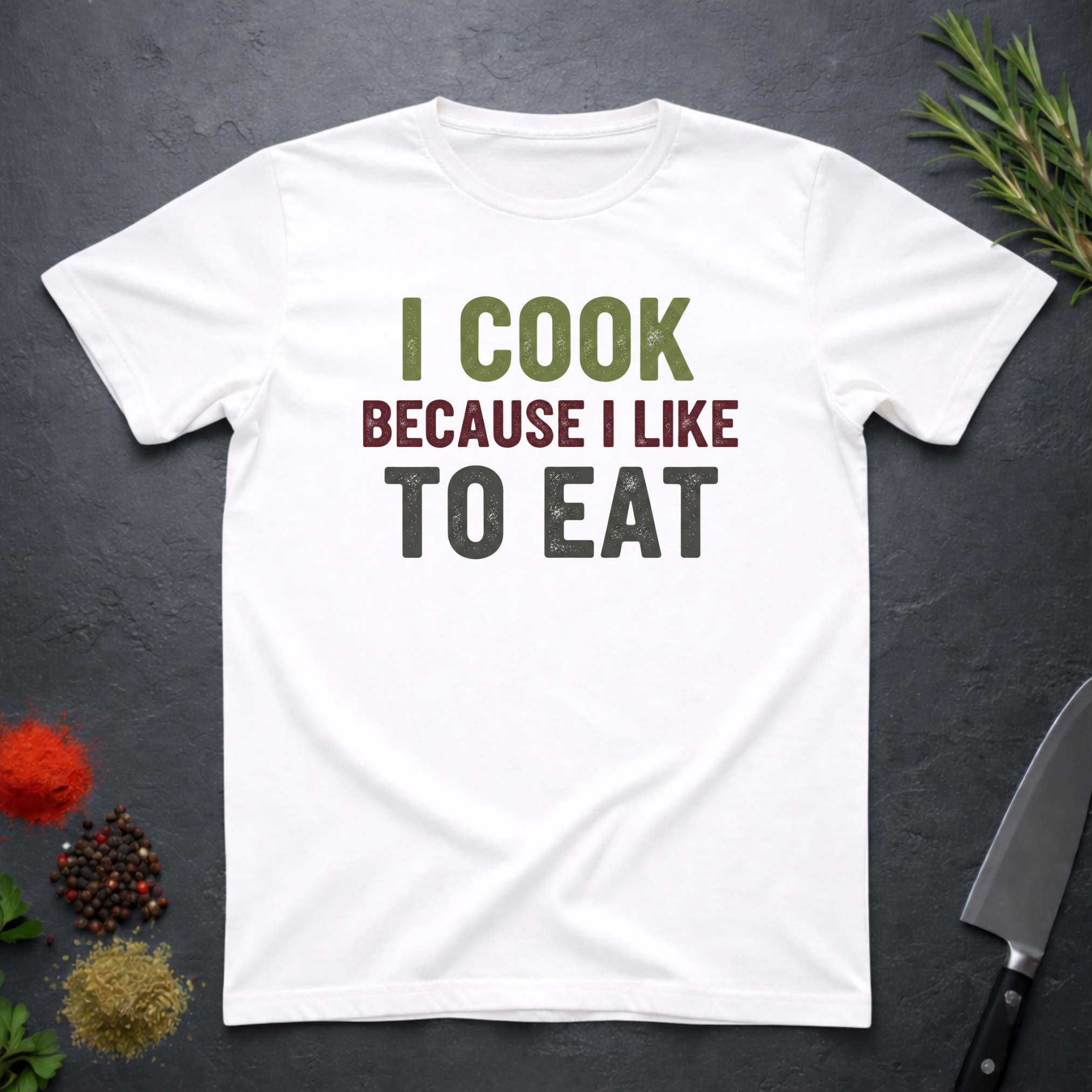 White t-shirt with 'I cook because I like to eat' text on a dark surface with kitchen items.