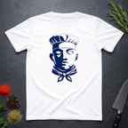 White t-shirt with dark blue graphic chef face design on a dark surface with kitchen items.