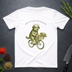 White t-shirt with a graphic of a dog riding a bicycle with a basket of vegetables and text 'off to feed the masses' on a dark surface.