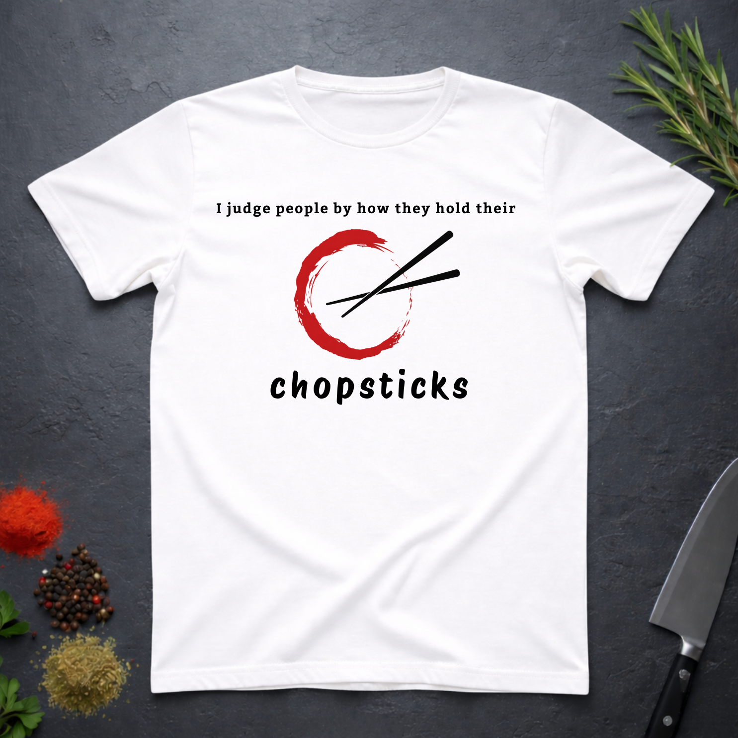 White t-shirt with Chopsticks graphic and text "I judge people by how they hold their chopsticks" on a dark surface with kitchen items.