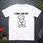 White t-shirt with a graphic design and text 'I COOK. YOU EAT.' on a dark surface with kitchen items.