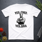 White t-shirt with a graphic of a chef and text 'YES, CHEF. YES, DAD.' on a dark surface with kitchen items.