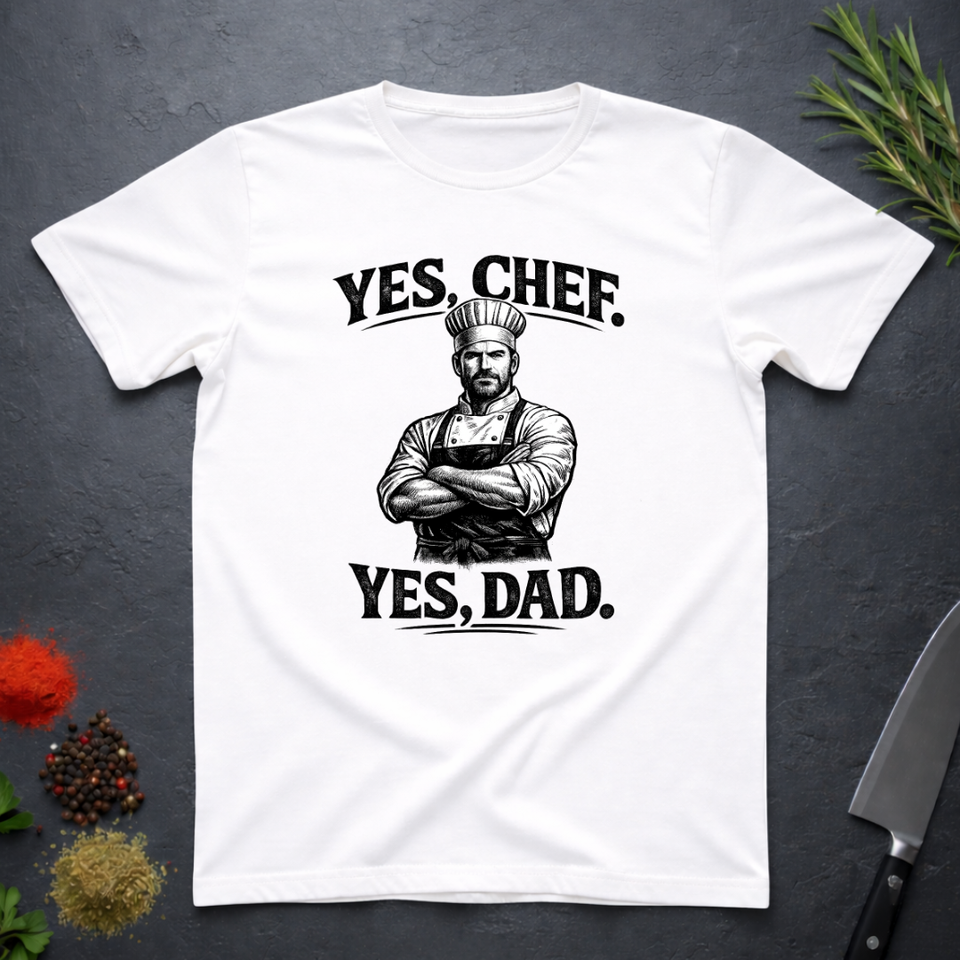 White t-shirt with a graphic of a chef and text 'YES, CHEF. YES, DAD.' on a dark surface with kitchen items.