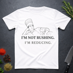 White t-shirt with a chef graphic and text 'I'm not rushing. I'm reducing' on a dark surface with kitchen items.