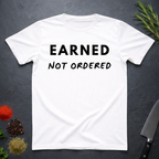 White t-shirt with 'Earned Not Ordered' text on a dark surface with decorative elements.