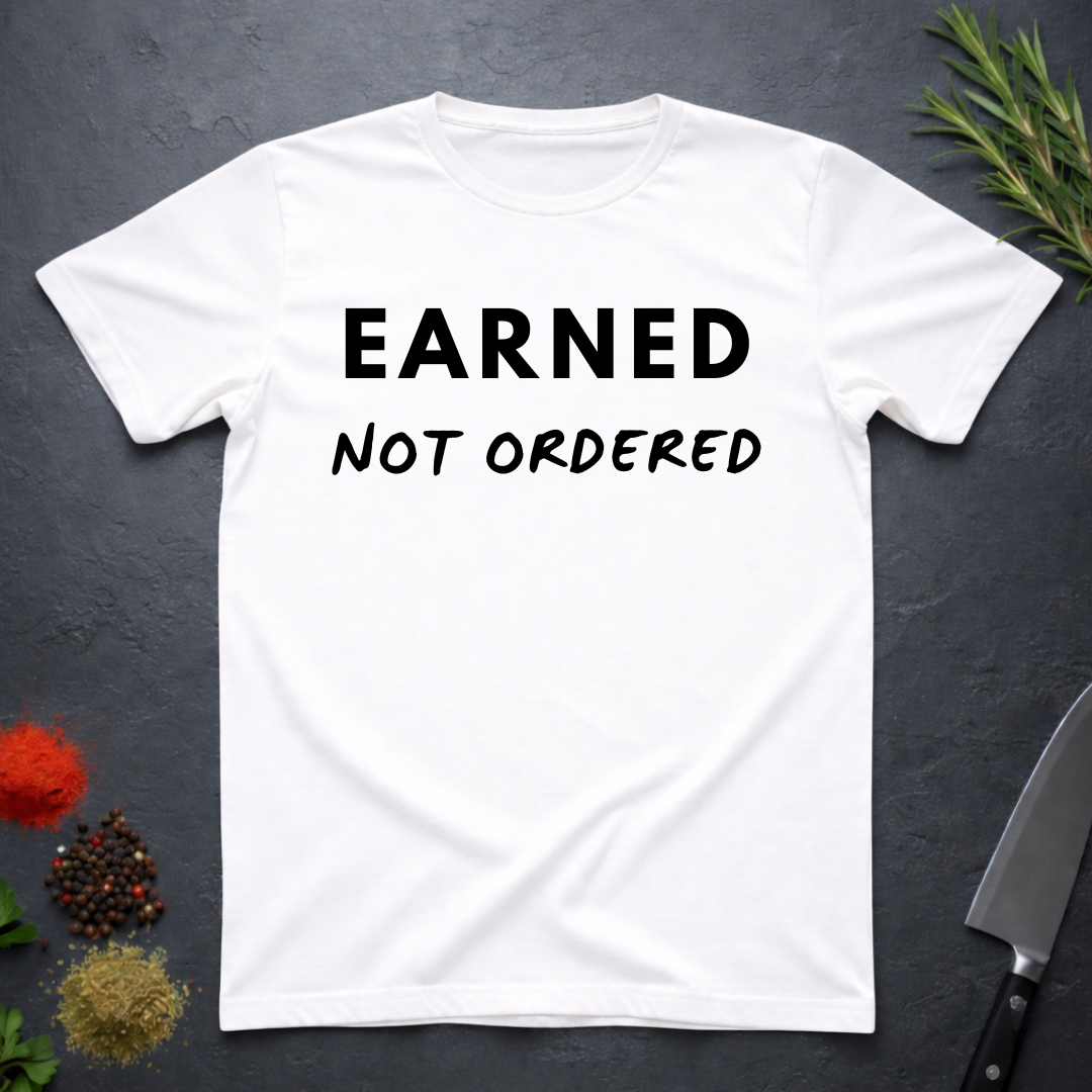White t-shirt with 'Earned Not Ordered' text on a dark surface with decorative elements.