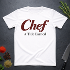 White t-shirt with 'Chef A Title Earned' text on a dark surface with kitchen items.