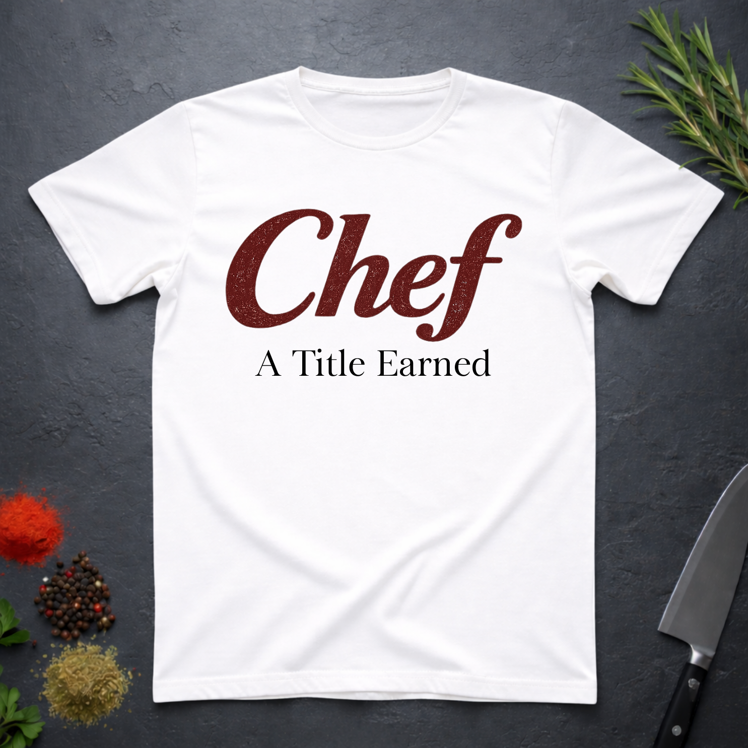 White t-shirt with 'Chef A Title Earned' text on a dark surface with kitchen items.