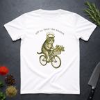White t-shirt with a cat riding a bicycle illustration and text 'off to feed the masses' on a dark surface.
